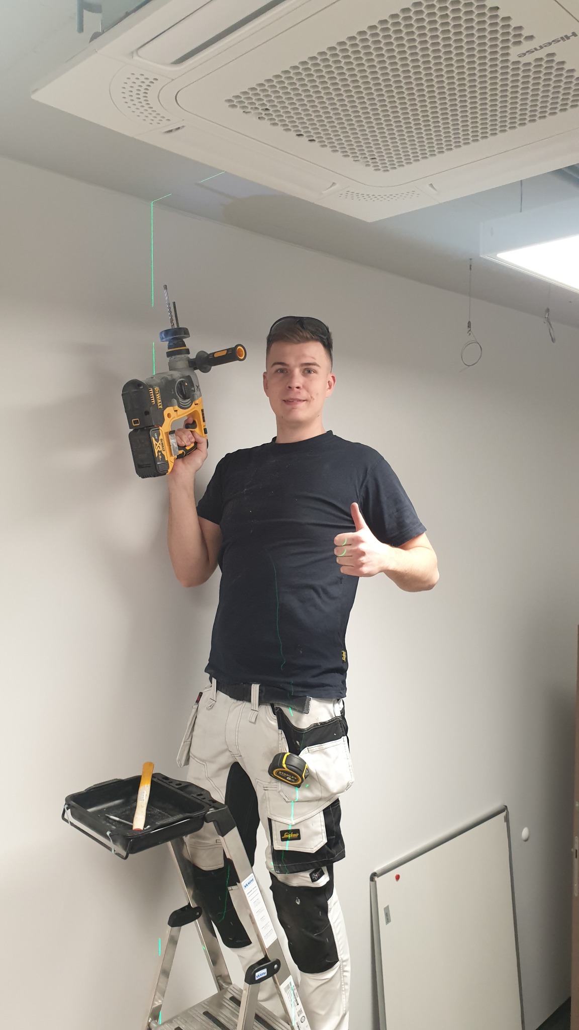 Uśmiechnięty monter na drabinie z wiertarką DeWalt z laserem krzyżowym celuje w sufit z klimatyzacją kasetonową, ubrany w robocze spodnie Snickers z narzędziami w kieszeniach i czarną koszulkę.