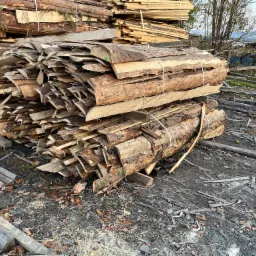Stosy świeżo przetartej tarcicy z korą, spięte sznurkiem, ułożone na placu tartacznym z widocznymi resztkami drewna i trocinami, w tle drzewa i zabudowania.