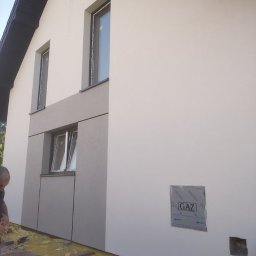 TermoHouse usługi remontowo budowlane Patryk Goleń - Dom w trakcie prac elewacyjnych: fragment ściany z oknami, częściowo pokryty szarymi panelami, widoczna skrzynka gazowa, zabezpieczona folia na ziemi i pracownik w lewym dolnym rogu.
