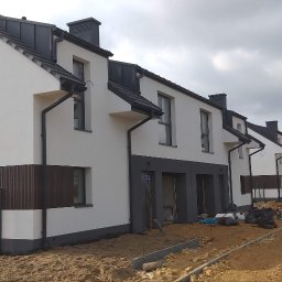 TermoHouse usługi remontowo budowlane Patryk Goleń - Szereg nowych domów w trakcie prac wykończeniowych elewacji, z widocznym jasnym tynkiem, ciemnymi elementami dekoracyjnymi i sprzętem budowlanym na placu budowy.