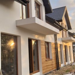 TermoHouse usługi remontowo budowlane Patryk Goleń - Nowoczesny dom jednorodzinny z elementami elewacji imitującymi drewno, białe ściany, brązowe okna, widoczne worki z zaprawą i drabina, teren wokół w trakcie prac wykończeniowych.