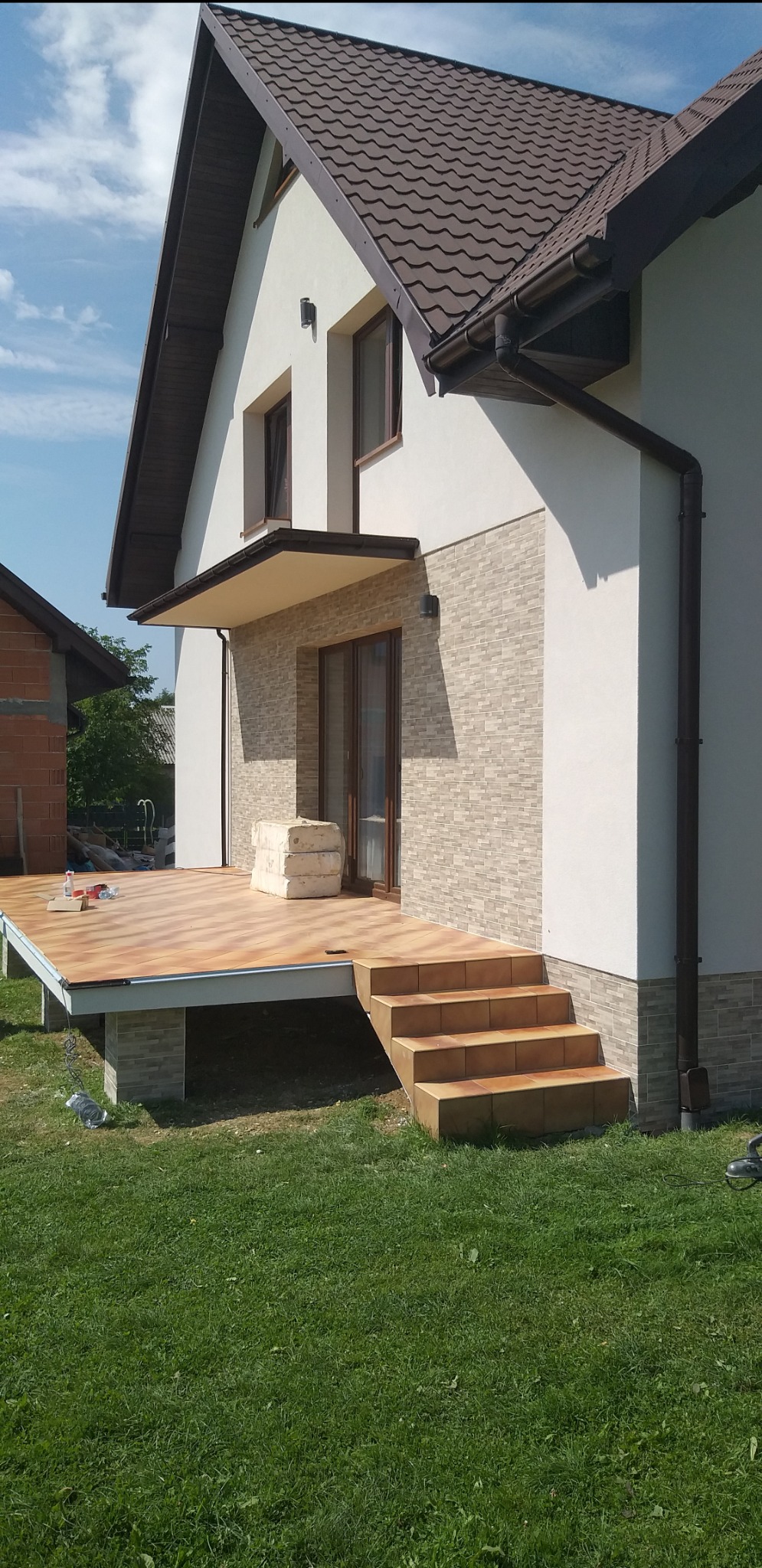Boczny widok domu z nową elewacją z jasnej cegły i drewnianym tarasem z brązowymi płytkami, schody z płytek prowadzą na trawnik.