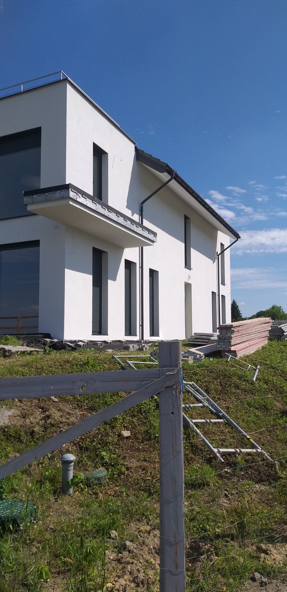 Nowoczesny, biały dom jednorodzinny z szarymi oknami i dachem, widoczny z dołu w słoneczny dzień, z elementami rusztowania i materiałami budowlanymi wokół, sugerującymi trwające prace wykończeniowe.