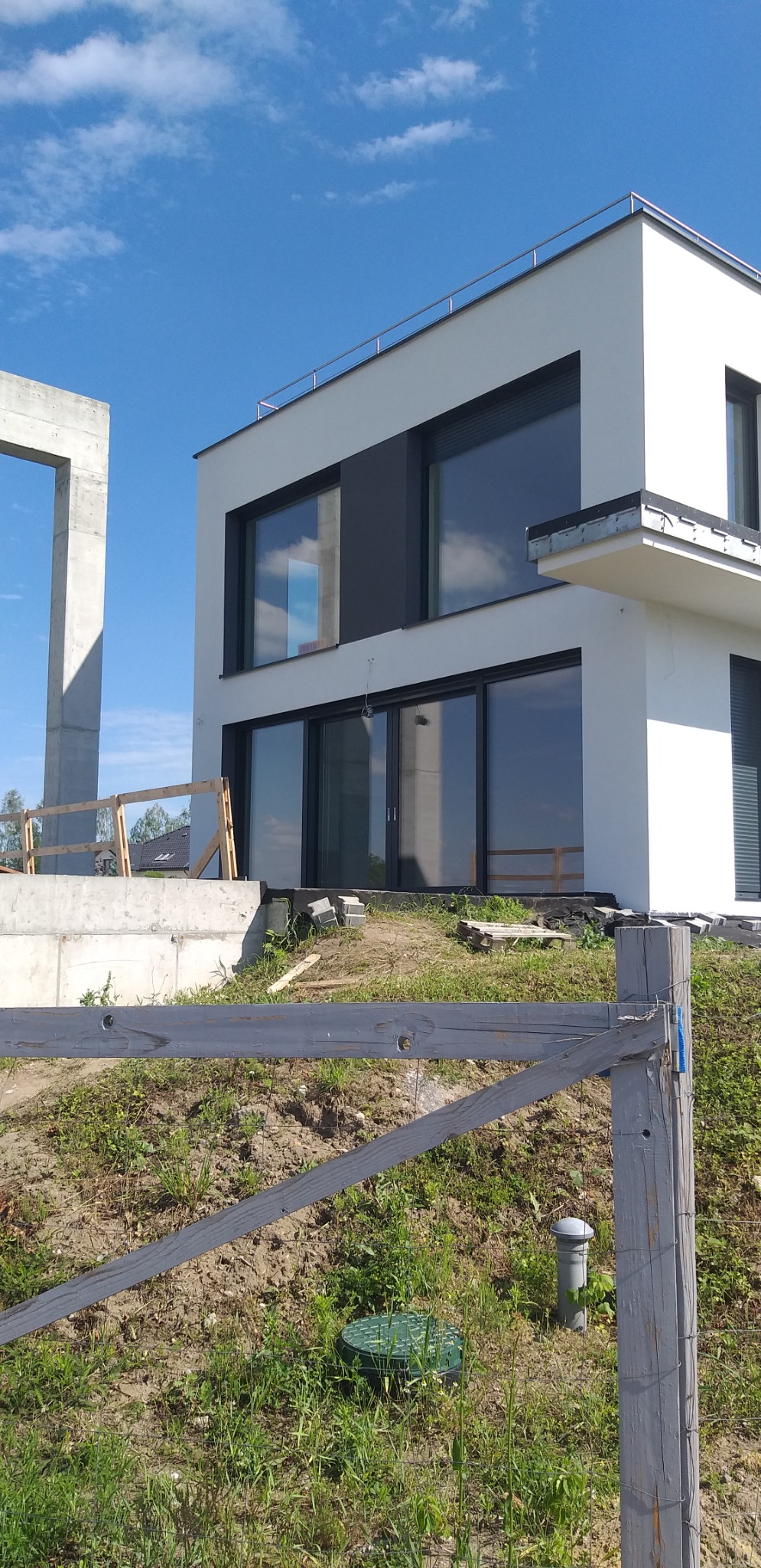 Nowoczesny, dwukondygnacyjny dom z białymi elewacjami i dużymi oknami, w trakcie wykończenia terenu wokół budynku; widoczny fragment betonowej konstrukcji i drewniane ogrodzenie.