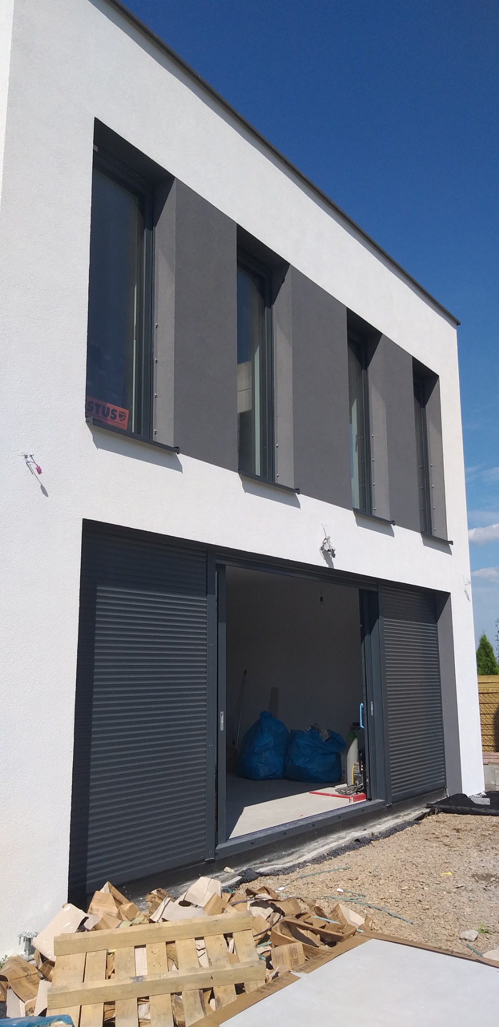 Nowoczesny dom z białą elewacją i szarymi elementami okiennymi, otwartymi roletami garażowymi, widoczne materiały budowlane na zewnątrz.