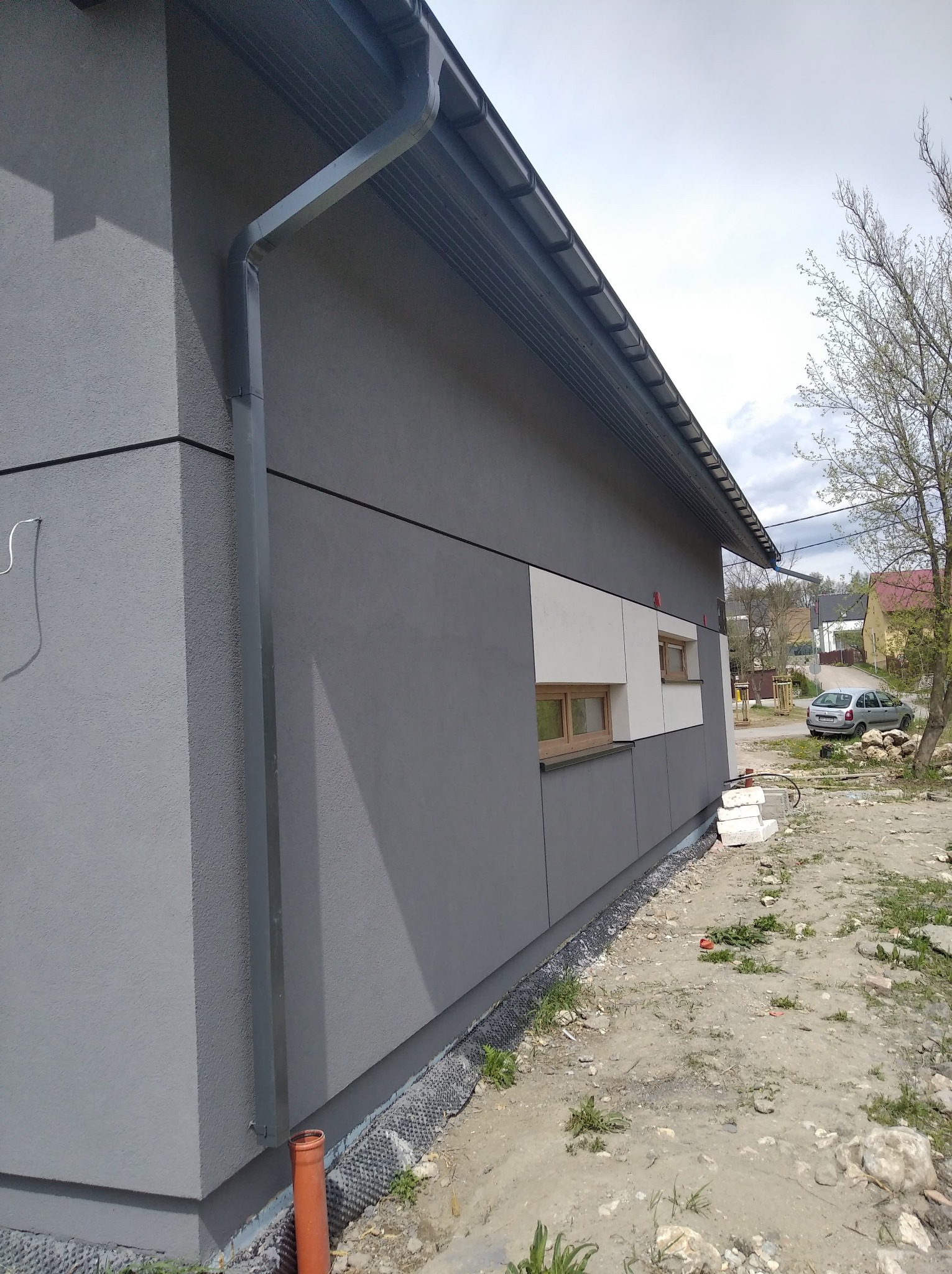 Szara elewacja budynku z drewnianymi oknami i systemem rynnowym, widoczne białe panele elewacyjne i teren wokół budynku.
