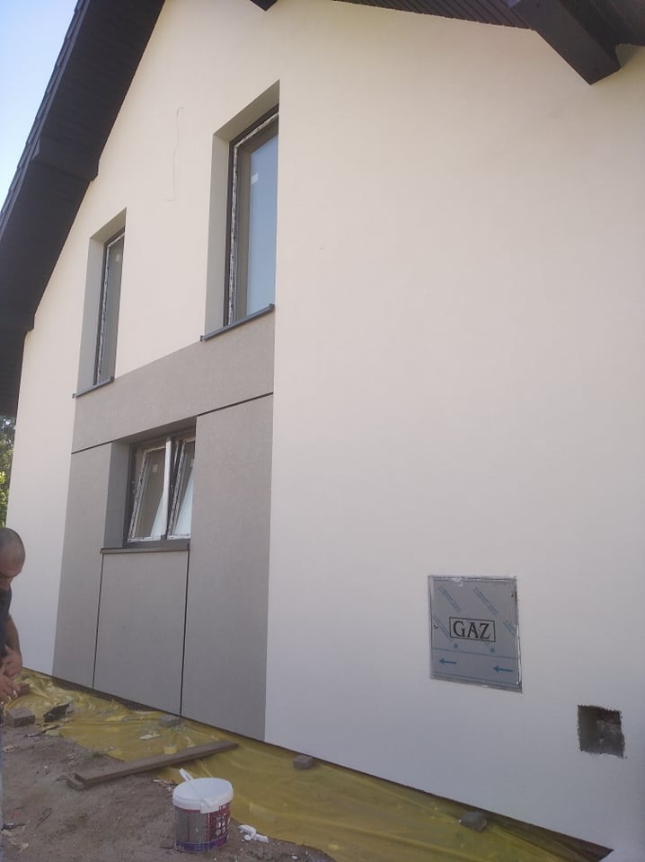 Dom w trakcie prac elewacyjnych: fragment ściany z oknami, częściowo pokryty szarymi panelami, widoczna skrzynka gazowa, zabezpieczona folia na ziemi i pracownik w lewym dolnym rogu.