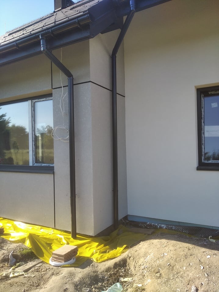 Fragment elewacji budynku z oknami i rynnami, zabezpieczony żółtą folią ochronną na gruncie.