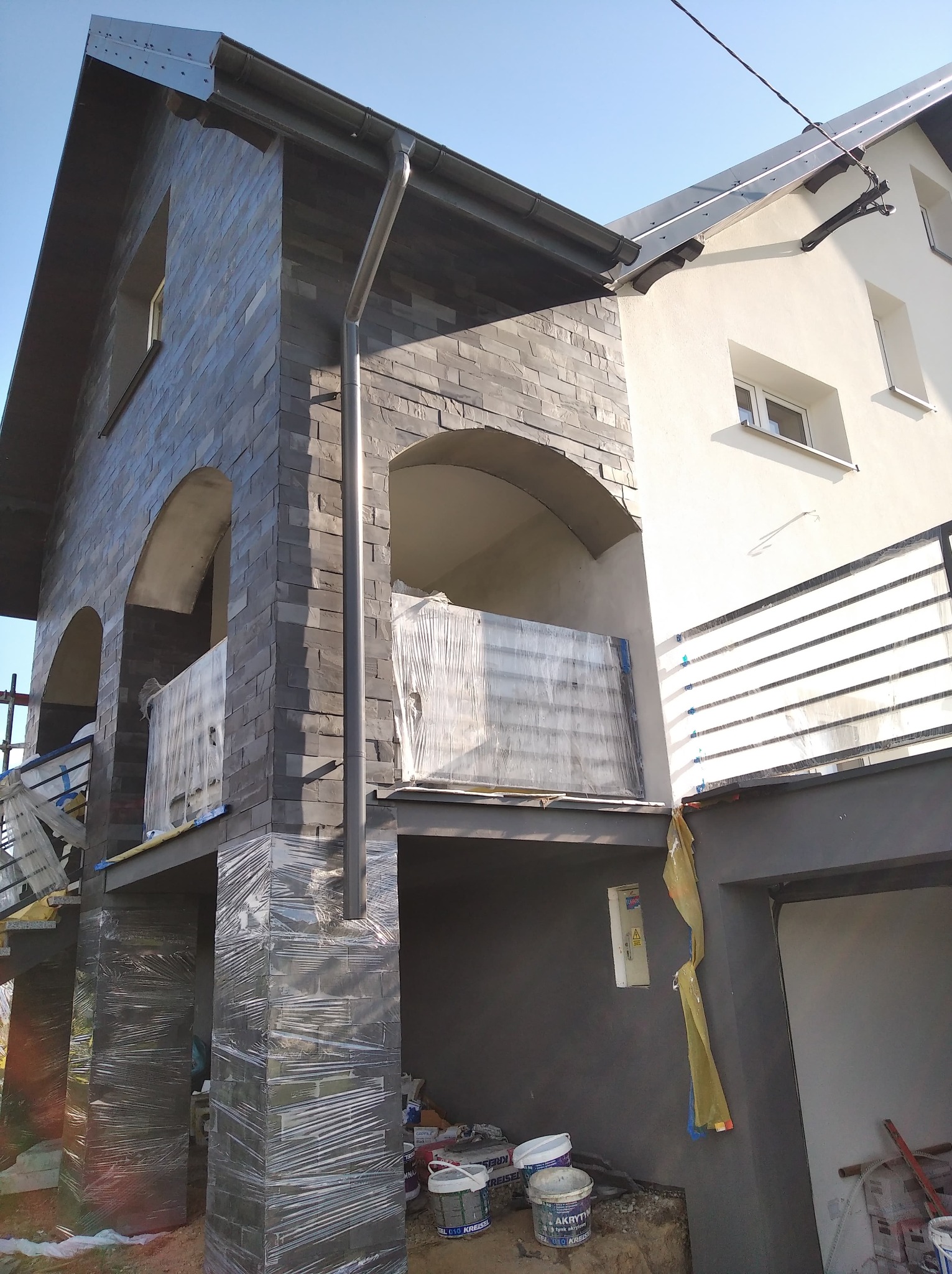 Dom w trakcie wykańczania elewacji z ciemnoszarej imitacji kamienia, zabezpieczony folią, z widocznymi puszkami farb i narzędziami na ziemi.