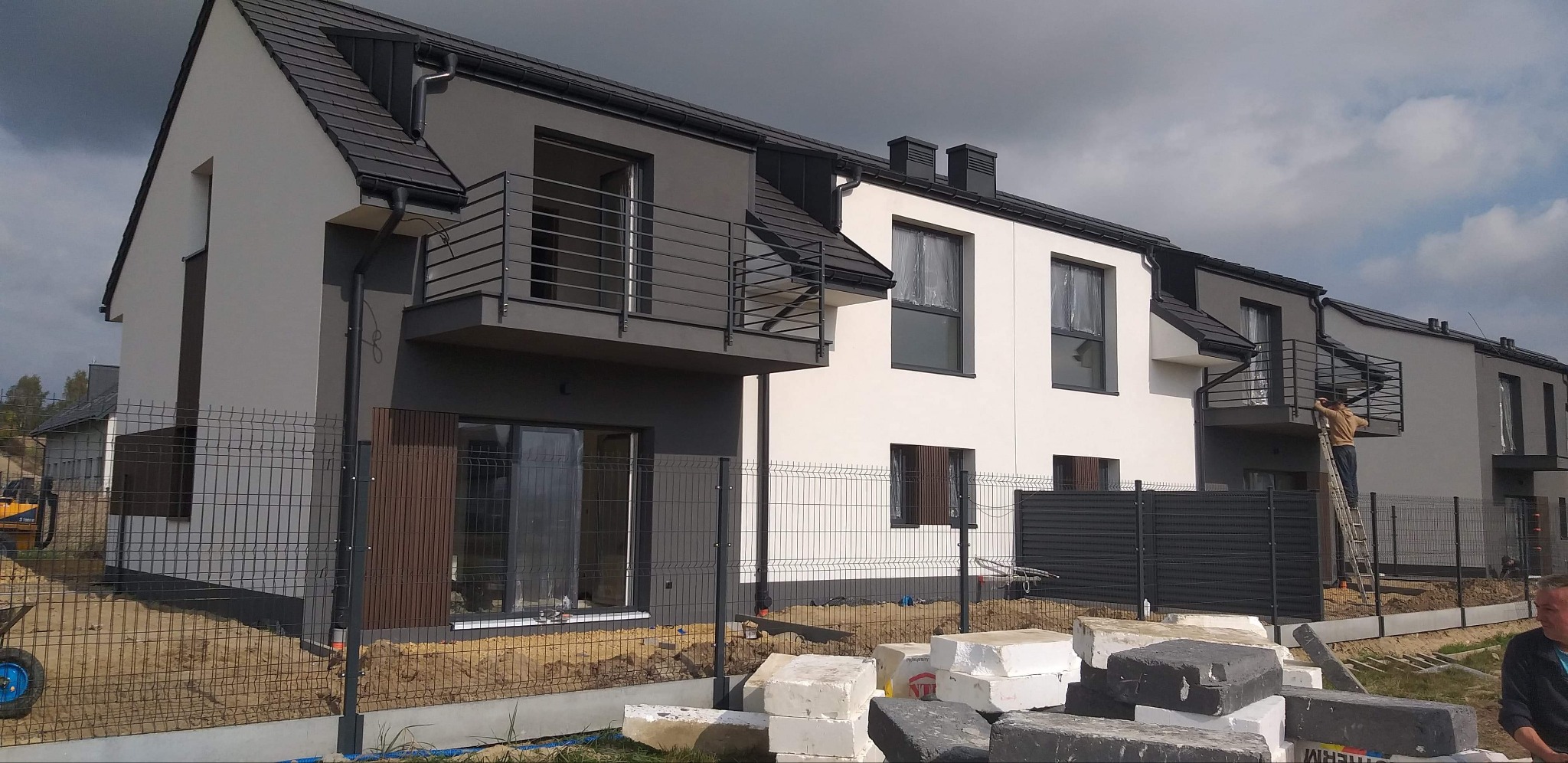 Nowo wybudowane domy w trakcie wykańczania elewacji, widoczne rusztowania i materiały budowlane, jeden z pracowników na drabinie na balkonie.