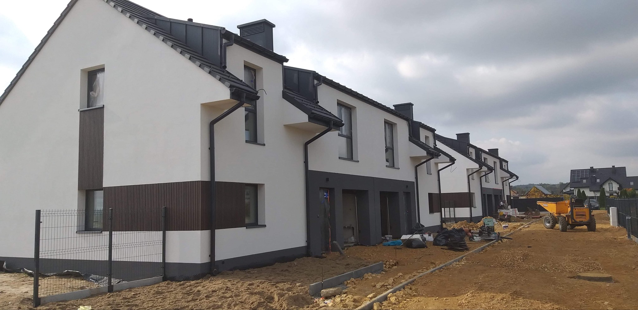Szereg nowych domów w trakcie prac wykończeniowych elewacji, z widocznym jasnym tynkiem, ciemnymi elementami dekoracyjnymi i sprzętem budowlanym na placu budowy.