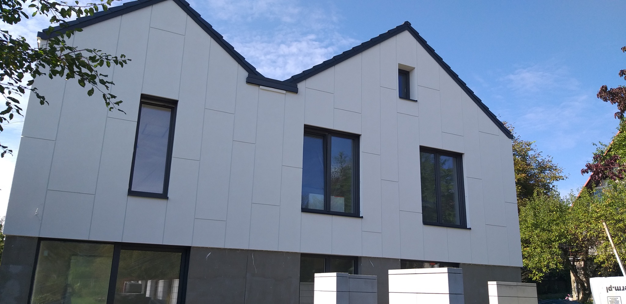 Nowoczesny dom z białą elewacją panelową i czarną stolarką okienną, widoczny z zewnątrz podczas słonecznego dnia.