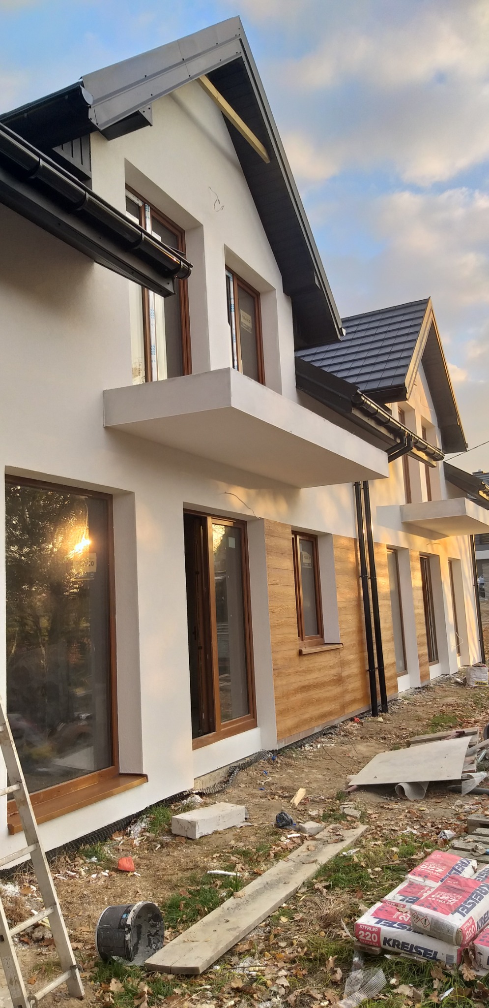 Nowoczesny dom jednorodzinny z elementami elewacji imitującymi drewno, białe ściany, brązowe okna, widoczne worki z zaprawą i drabina, teren wokół w trakcie prac wykończeniowych.