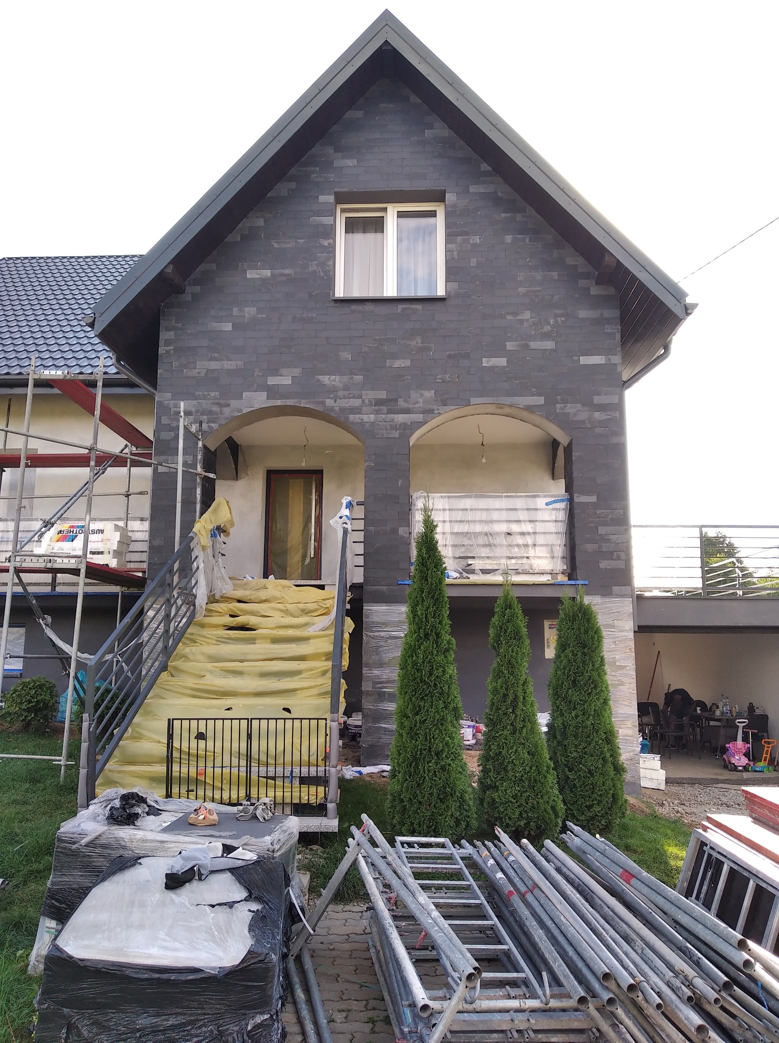 Dom z elewacją z szarej cegły klinkierowej w trakcie wykańczania, zabezpieczone schody folią, widoczne rusztowania i materiały budowlane.