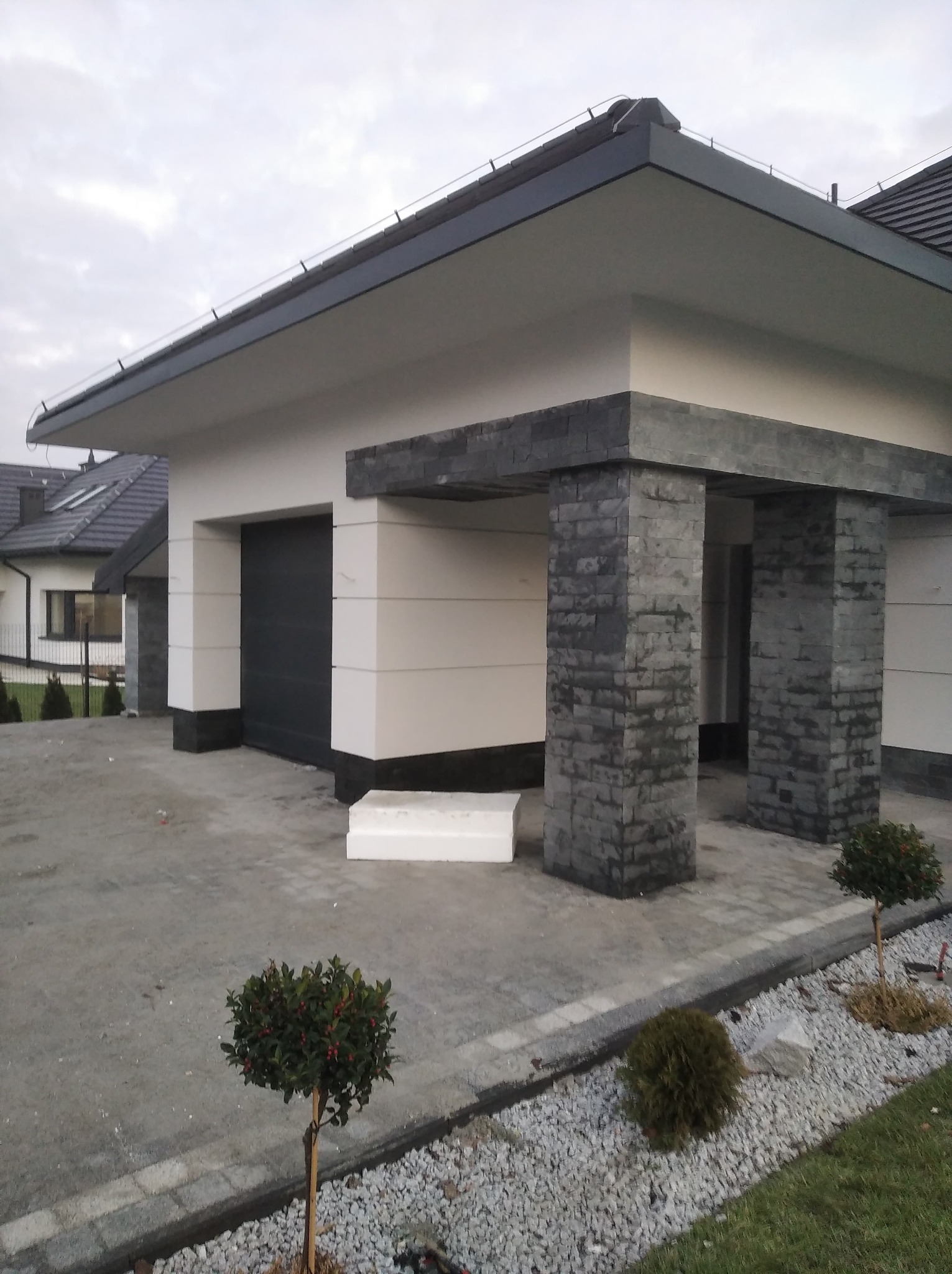 Nowoczesny dom z garażem i podjazdem wyłożonym kostką, z elementami dekoracyjnymi z kamienia na elewacji i kolumnach, małe drzewka ozdobne posadzone wzdłuż podjazdu.