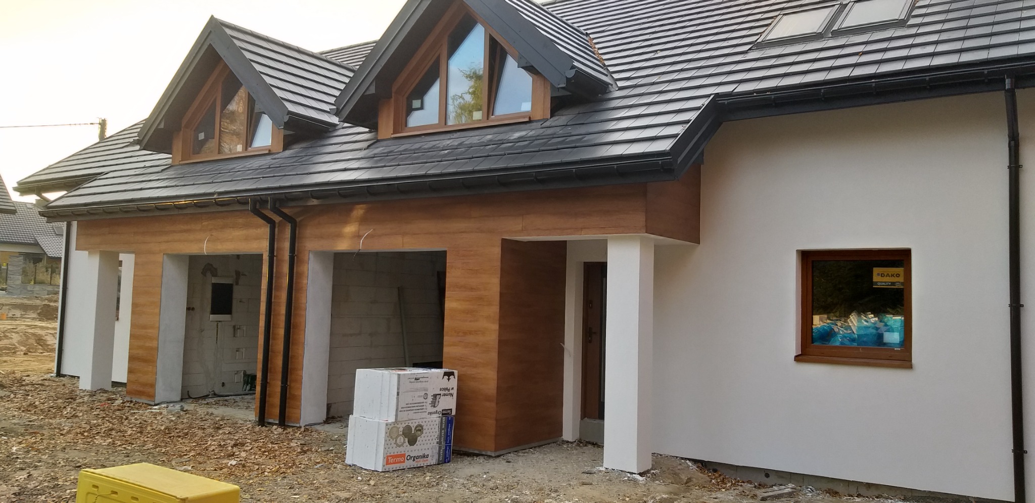 Nowoczesny dom z elewacją w kolorze drewna, widoczne okna dachowe i częściowo wykończony garaż, na ziemi leżą styropianowe płyty.