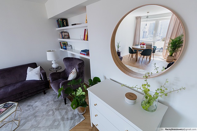 Nowoczesny salon z fioletową sofą i fotelem, białymi półkami na książki, okrągłym lustrem odbijającym jadalnię oraz białą komodą z roślinami.