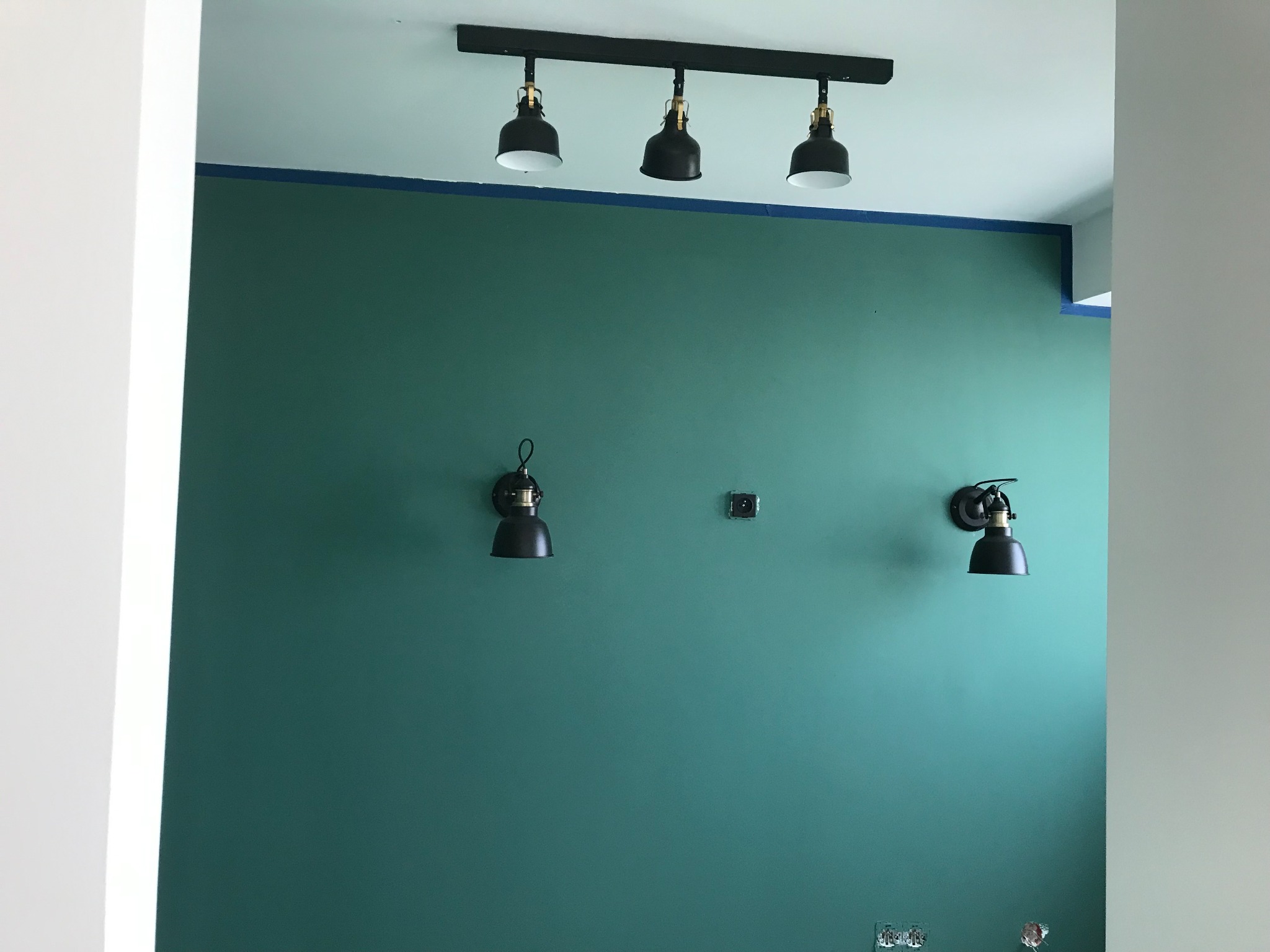 Ściana pomalowana na zielono-turkusowy kolor z trzema czarnymi reflektorami sufitowymi na szynie, dwoma kinkietami w stylu industrialnym i gniazdkiem elektrycznym.