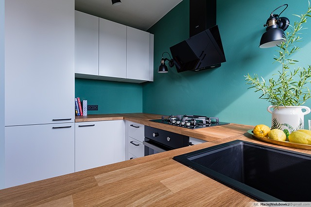 Nowoczesna kuchnia z białymi szafkami, drewnianym blatem, czarnym okapem i turkusową ścianą, oświetlona lampami w stylu industrialnym, z dekoracyjnym bukietem i cytrynami na blacie.