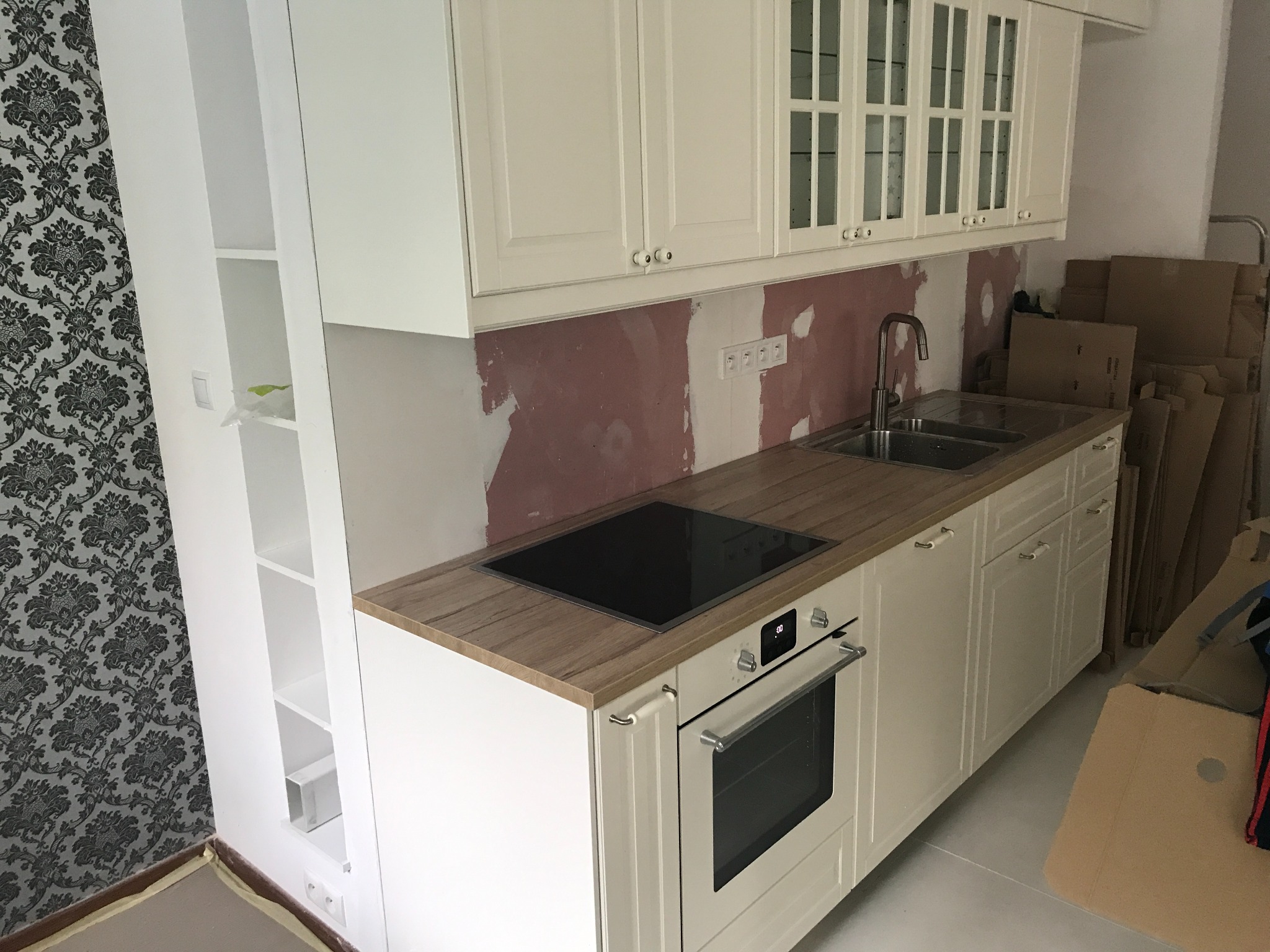 Wnętrze kuchni w trakcie remontu: białe szafki kuchenne z drewnianym blatem, płyta indukcyjna, piekarnik, ściana z widocznymi śladami po gładzi, tapeta w kwiaty, kartony w tle.