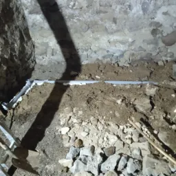 Fundament budynku w trakcie prac izolacyjnych: widoczne rury odprowadzające wodę, szpadle, drabina i kamienne ściany wykopu.