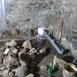 Wnętrze piwnicy z kamiennymi ścianami w trakcie prac instalacyjnych: widoczne rury kanalizacyjne, narzędzia i chemia gospodarcza. Otwór w ścianie z widocznym światłem z zewnątrz.