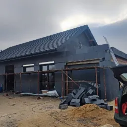 Dom jednorodzinny w trakcie prac związanych z izolacją elewacji, widoczne rusztowania, materiały izolacyjne i samochody dostawcze na posesji.