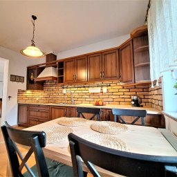 DOMITUS SPÓŁKA Z OGRANICZONĄ ODPOWIEDZIALNOŚCIĄ - Rustykalna kuchnia z ceglaną ścianą, drewnianymi szafkami i stołem z plecionymi podkładkami. Widok z perspektywy stołu, ciepłe oświetlenie.