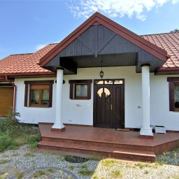 DOMITUS SPÓŁKA Z OGRANICZONĄ ODPOWIEDZIALNOŚCIĄ - Parterowy dom z brązowym dachem i drewnianą stolarką okienną, białą elewacją i kolumnami przy wejściu. Widok z zewnątrz, w tle zieleń drzew i błękitne niebo.