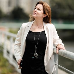 DOMITUS SPÓŁKA Z OGRANICZONĄ ODPOWIEDZIALNOŚCIĄ - Kobieta w białym żakiecie i czarnym stroju, oparta o barierkę, spogląda w górę na tle miejskiego krajobrazu z rozmytymi budynkami i zielenią.