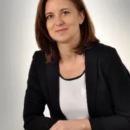 Portret kobiety w czarnej marynarce i białej bluzce, opierającej ręce na białym cokole z okularami, na neutralnym tle.