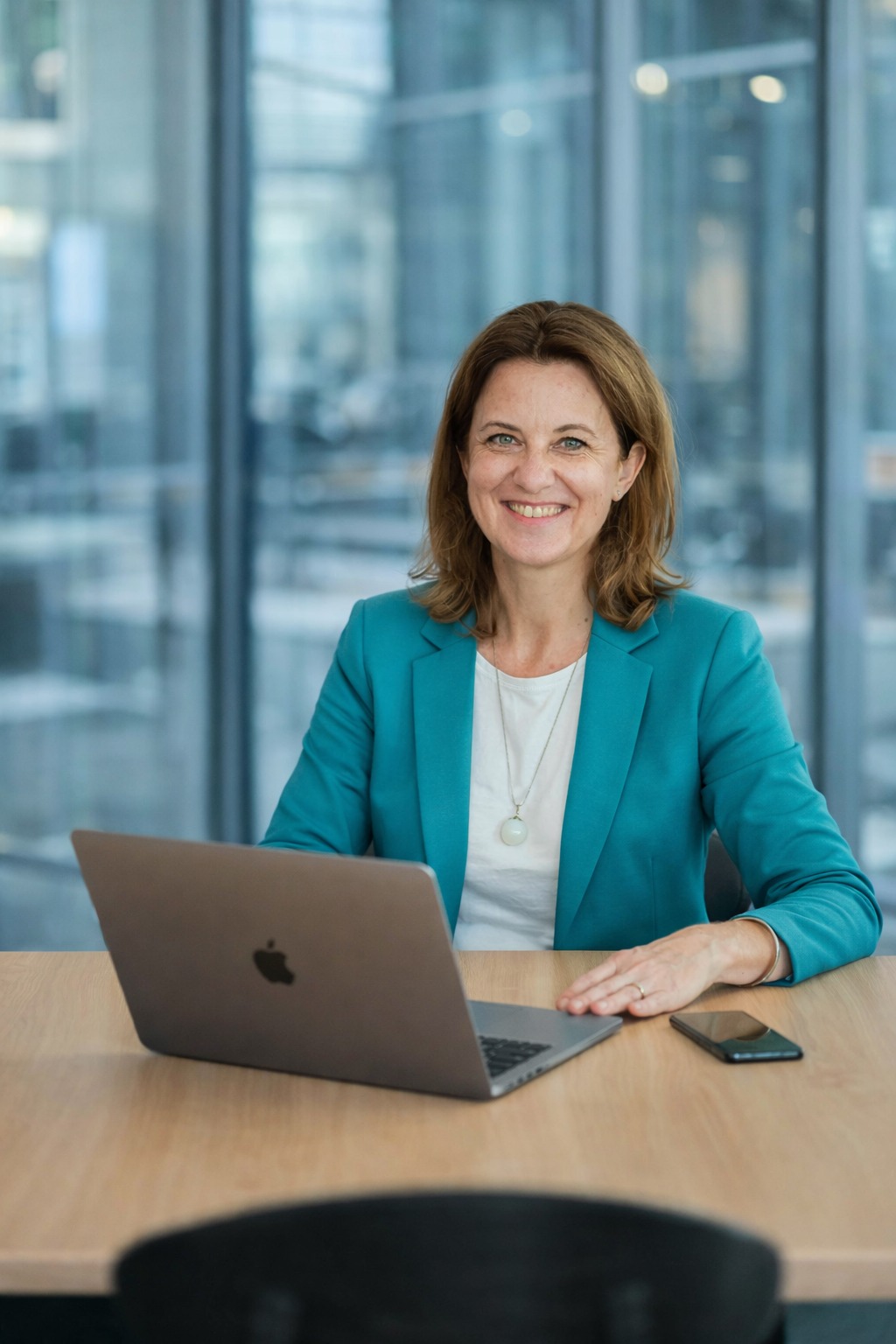 Uśmiechnięta kobieta w turkusowej marynarce siedzi za biurkiem z laptopem i telefonem. W tle przeszklona ściana biurowa.