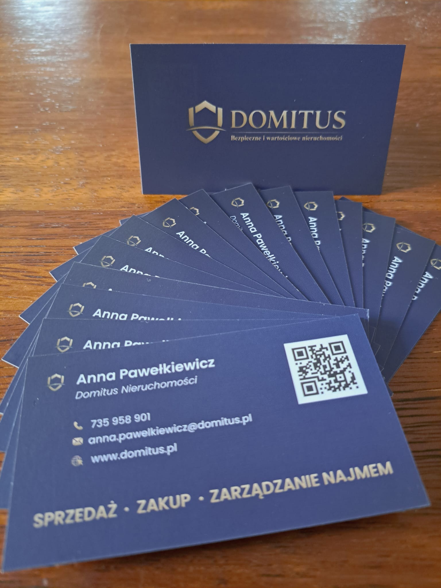 Wizytówki Domitus Nieruchomości w odcieniach granatu i złota, ułożone wachlarzowo, z widocznym logo i danymi kontaktowymi Anny Pawełkiewicz oraz hasłami: Sprzedaż, Zakup, Zarządzanie Najmem.