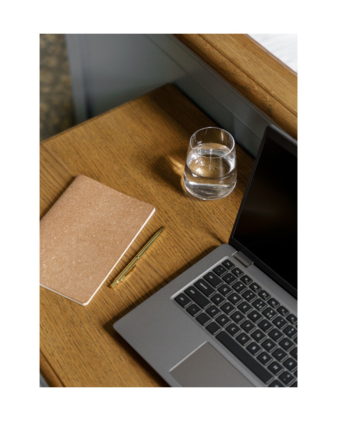 Laptop, notatnik, długopis i szklanka wody na drewnianym biurku w neutralnej tonacji, sugerujące pracę zdalną lub home office.
