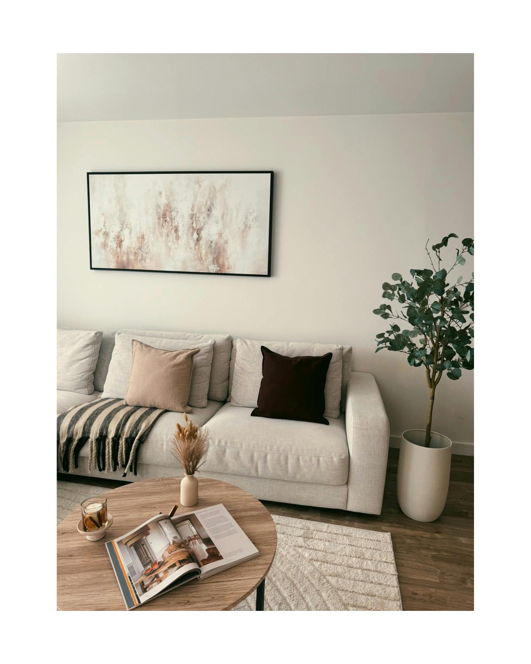 Jasne wnętrze salonu z beżową sofą, okrągłym stolikiem kawowym i abstrakcyjnym obrazem w czarnej ramie. Minimalistyczny wystrój z rośliną doniczkową i dywanem.