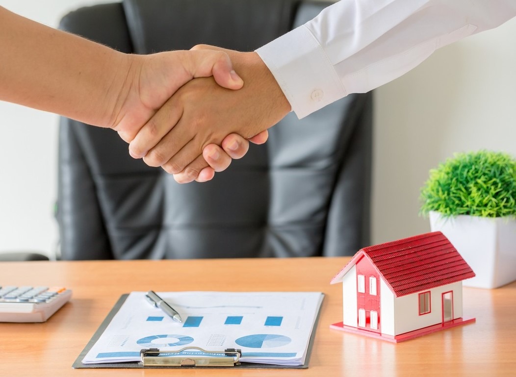 Uścisk dłoni nad stołem z modelem domu, kalkulatorem i dokumentami. Symbol zgody w transakcji nieruchomości.