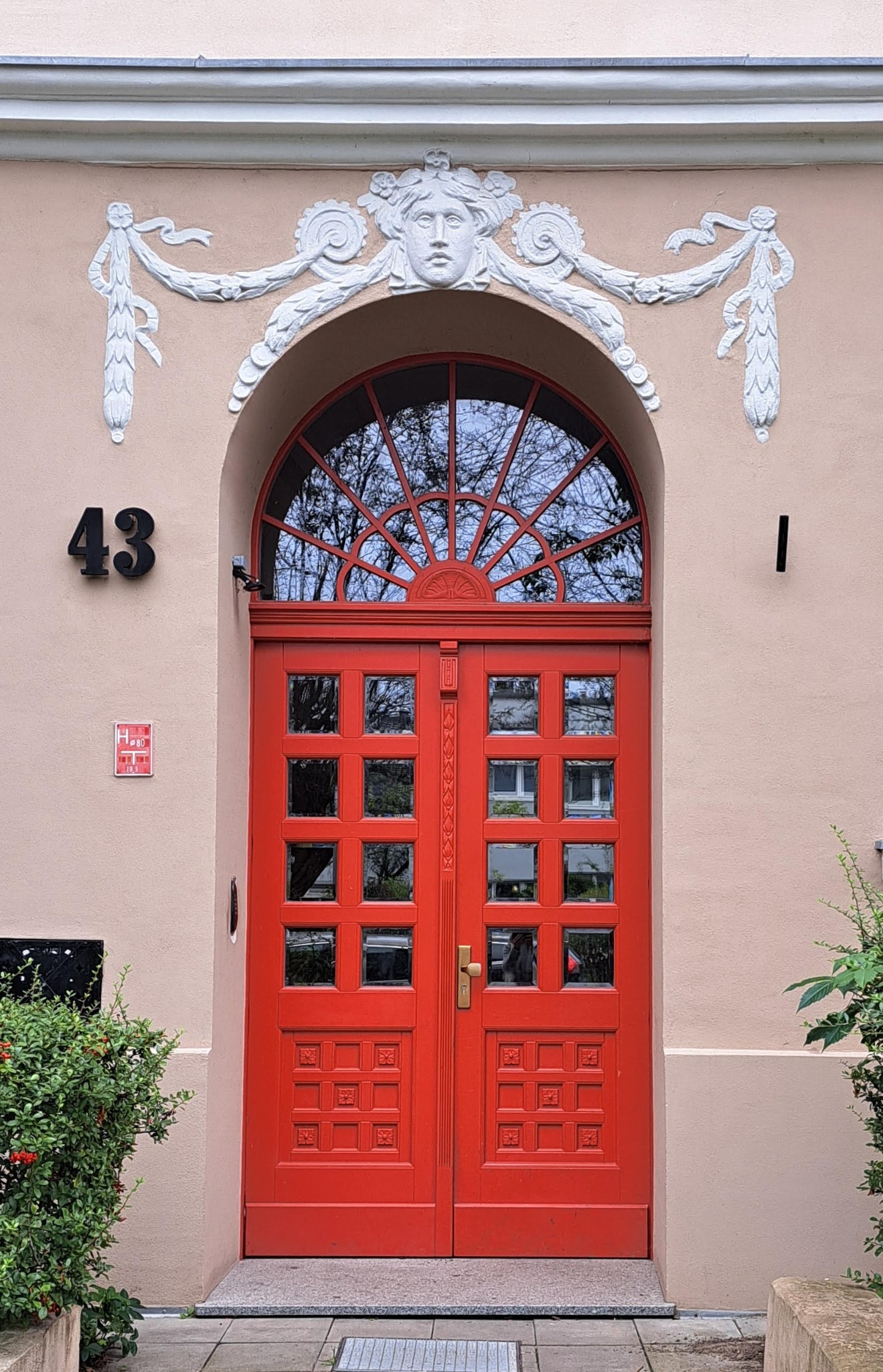 Elewacja budynku z czerwonymi, dwuskrzydłowymi drzwiami wejściowymi, numerem '43' i dekoracyjnym, białym ornamentem nad wejściem.