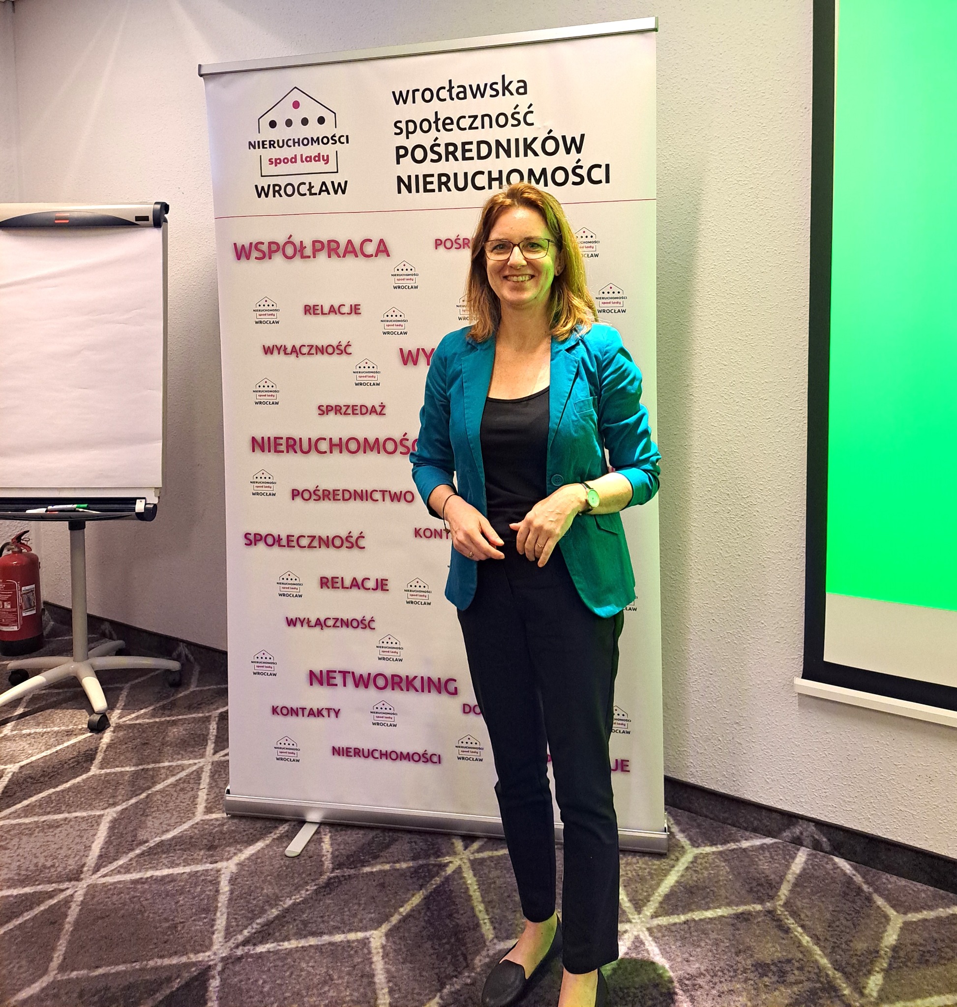 Kobieta w turkusowej marynarce pozuje na tle roll-upu z logo 'Nieruchomości spod lady Wrocław', prezentując ofertę agencji nieruchomości.