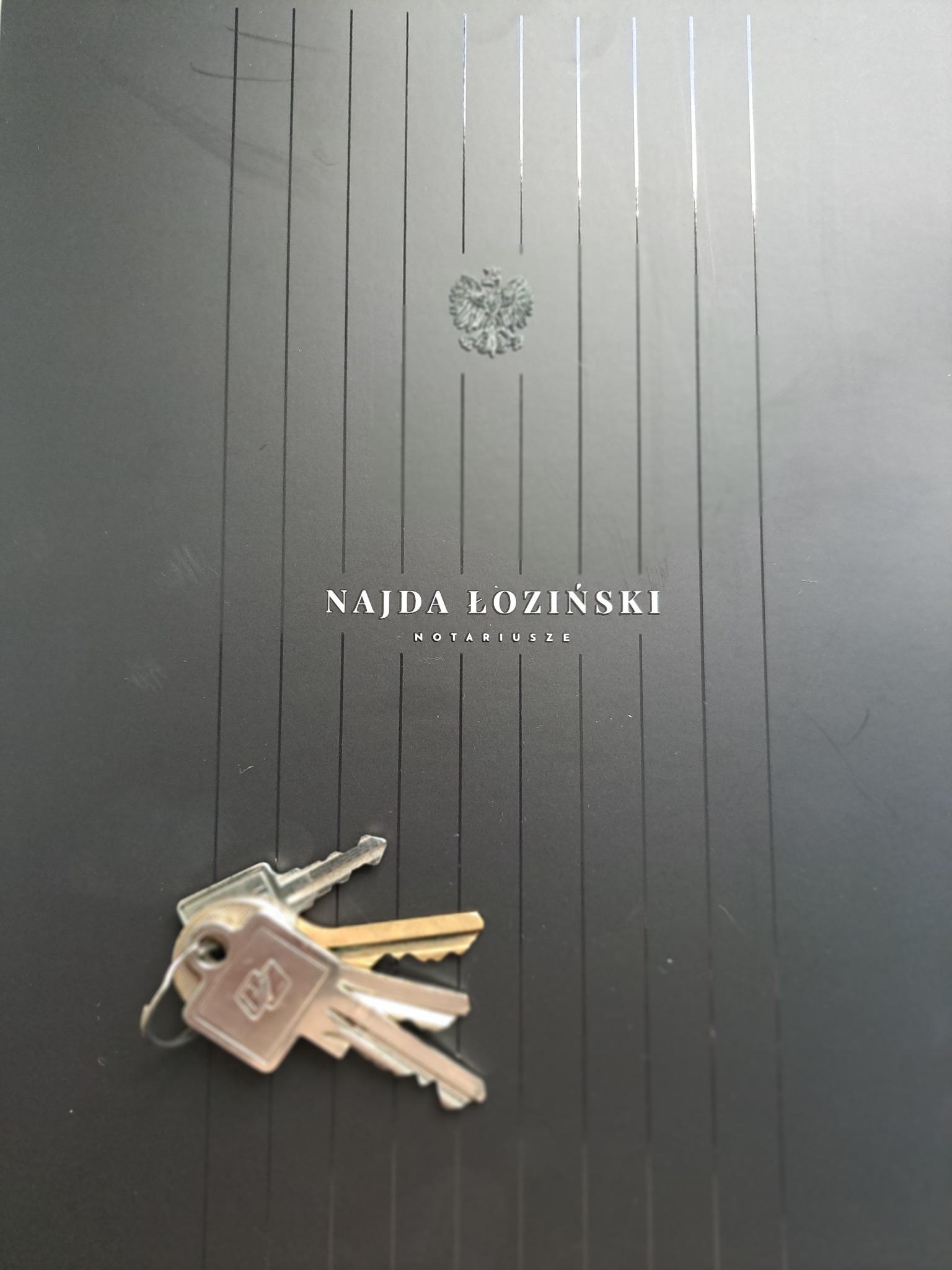 Klucze na czarnym folderze z godłem i napisem 'Najda Łoziński Notariusze'. Folder z pionowymi paskami. Ujęcie z góry, lekko z ukosa.