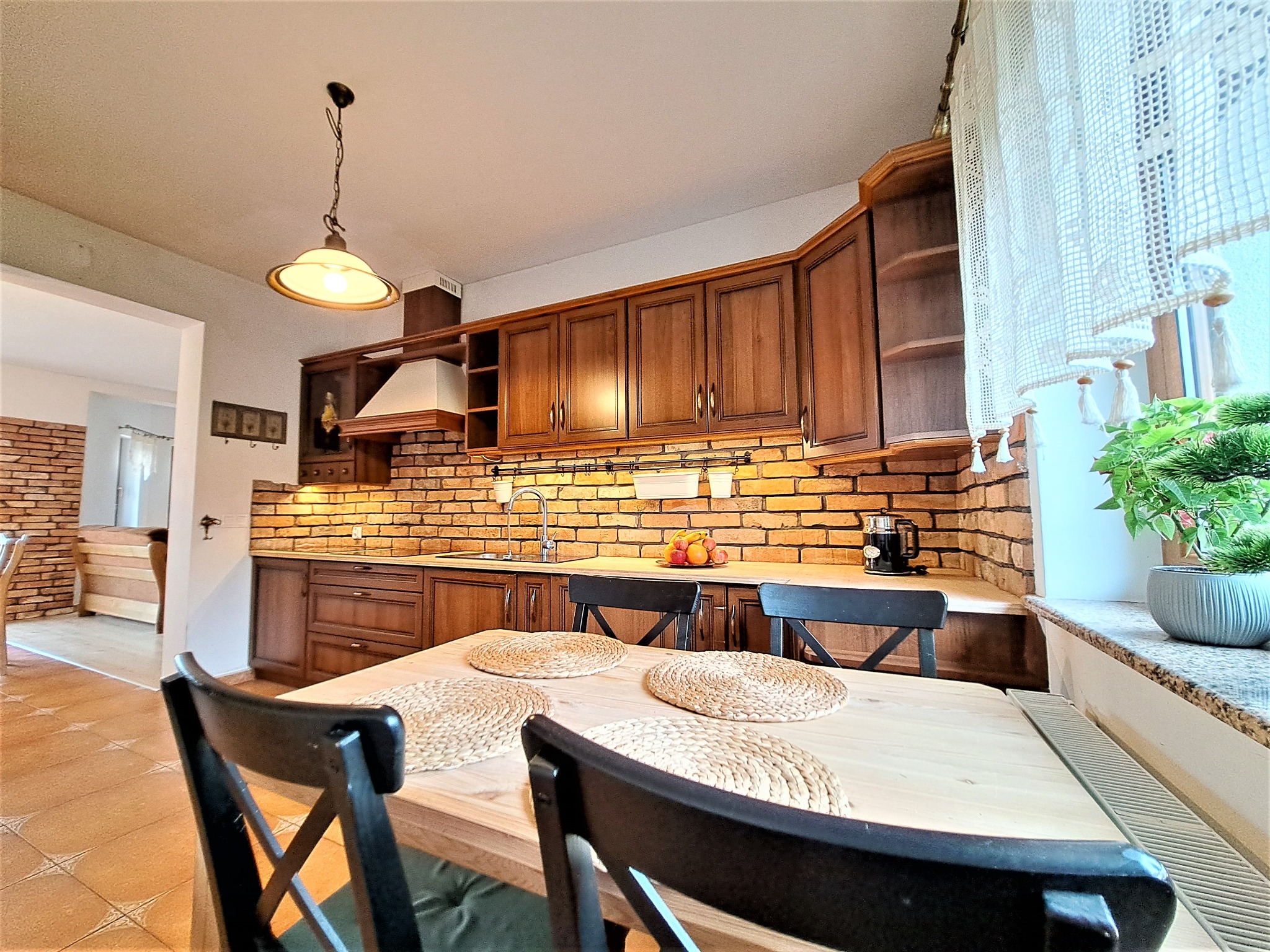 Rustykalna kuchnia z ceglaną ścianą, drewnianymi szafkami i stołem z plecionymi podkładkami. Widok z perspektywy stołu, ciepłe oświetlenie.