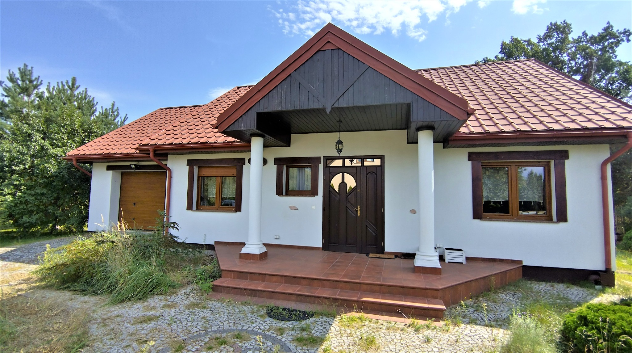 Parterowy dom z brązowym dachem i drewnianą stolarką okienną, białą elewacją i kolumnami przy wejściu. Widok z zewnątrz, w tle zieleń drzew i błękitne niebo.