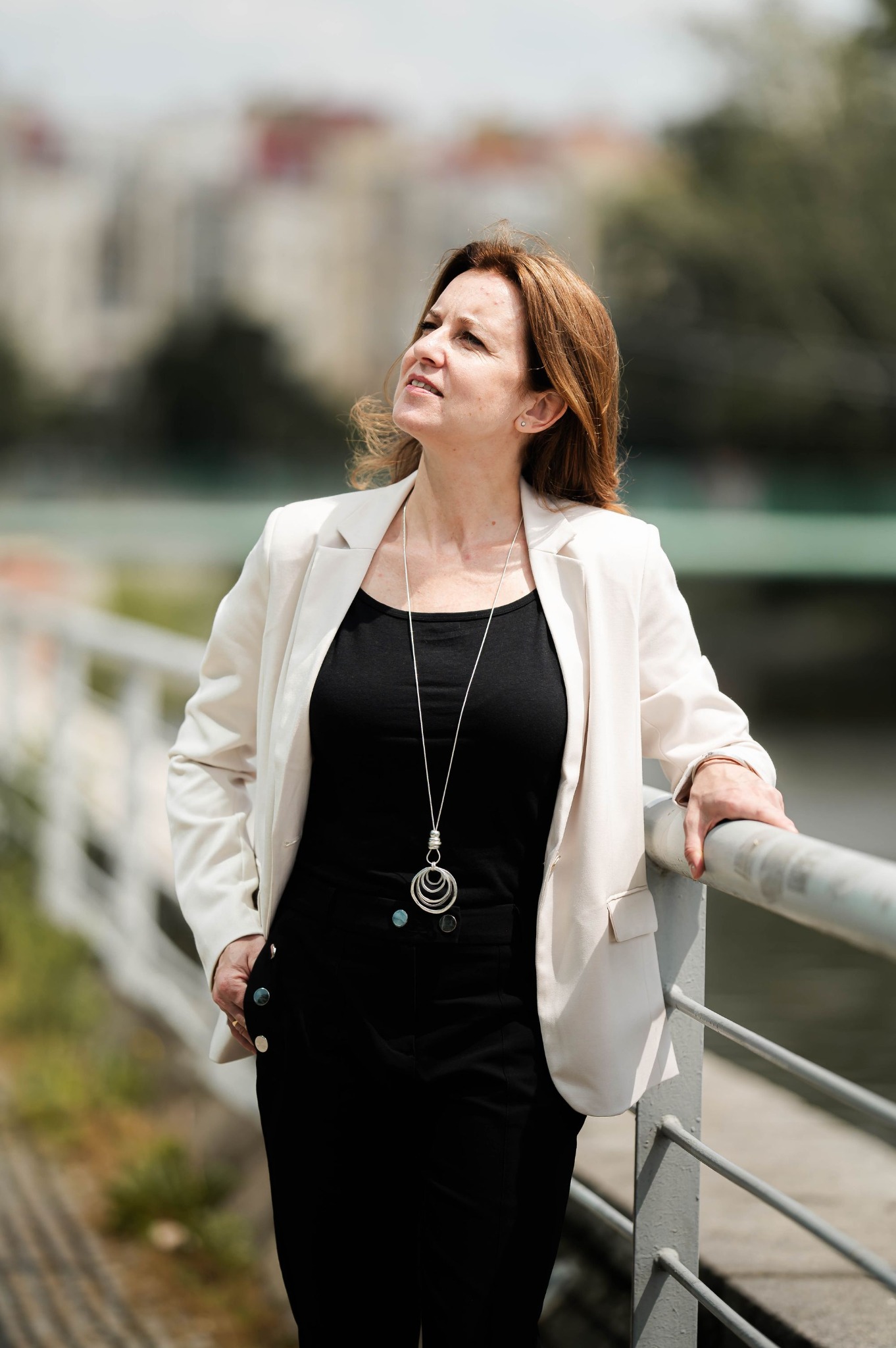 Kobieta w białym żakiecie i czarnym stroju, oparta o barierkę, spogląda w górę na tle miejskiego krajobrazu z rozmytymi budynkami i zielenią.