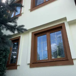 Elewacja budynku po ociepleniu, z widocznymi oknami w brązowych ramach i strukturą tynku baranka, częściowo zasłonięta przez gałąź świerku