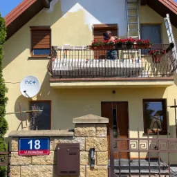 Mężczyzna maluje fragment elewacji domu z balkonu, widoczne rusztowanie i drabiny, częściowo pomalowana ściana szczytowa, donice z czerwonymi kwiatami na balustradzie.