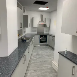 Jasna kuchnia z szarymi szafkami i blatem z brokatem, widoczny zlewozmywak, płyta grzewcza i okap, podłoga wyłożona panelami imitującymi drewno, widoczne ślady po remoncie.