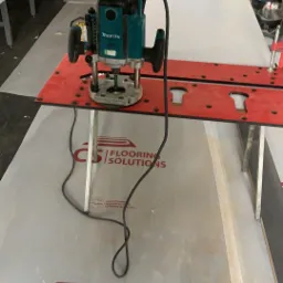 Frezarka Makita na czerwonym szablonie do wycinania otworów w panelach podłogowych, widoczne logo 'CS Flooring Solutions' na podkładzie pod panele.