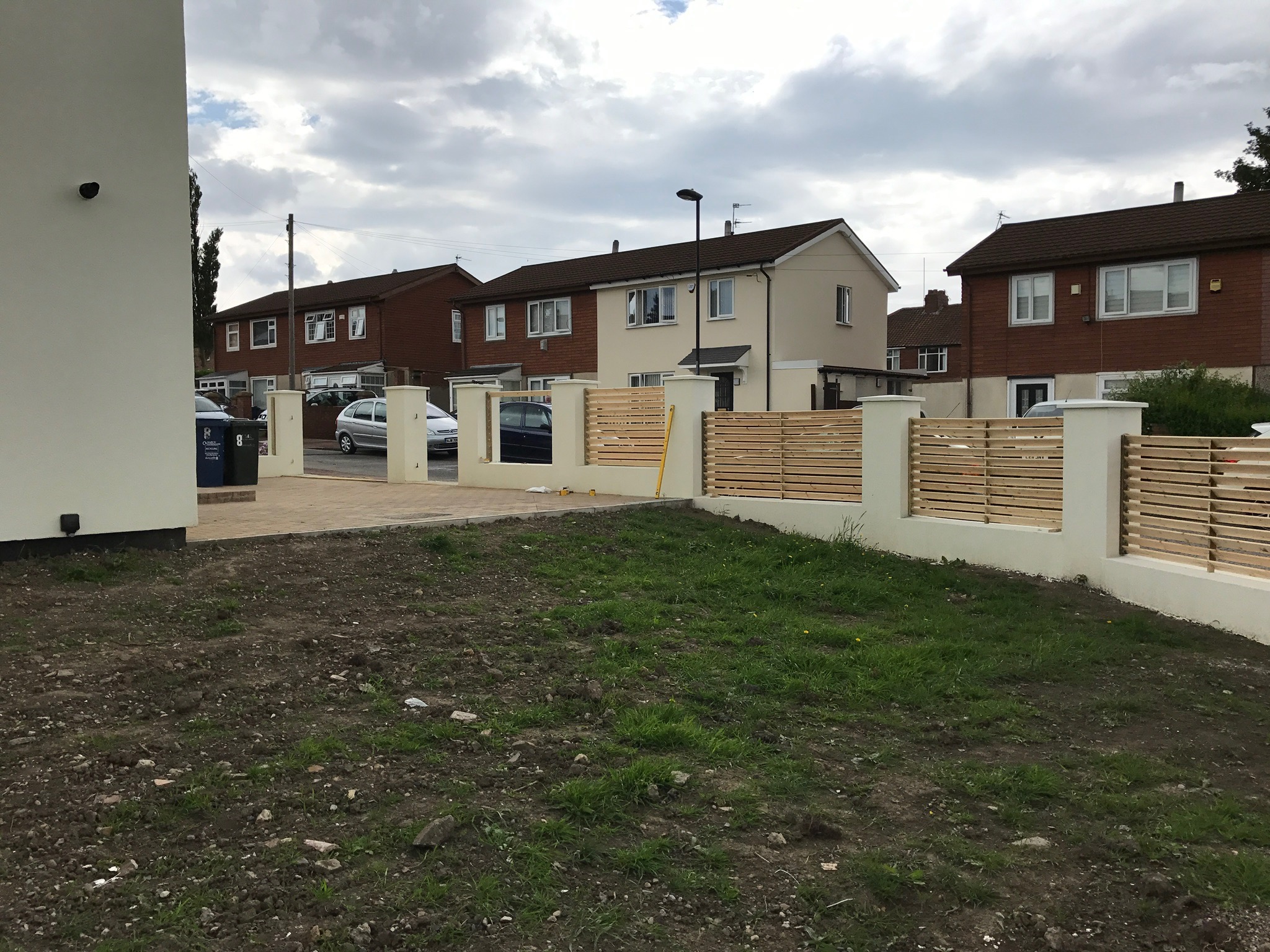 Nowoczesne ogrodzenie posesji z poziomych drewnianych listew, osadzone na białych, murowanych słupkach, na tle domów jednorodzinnych w pochmurny dzień.