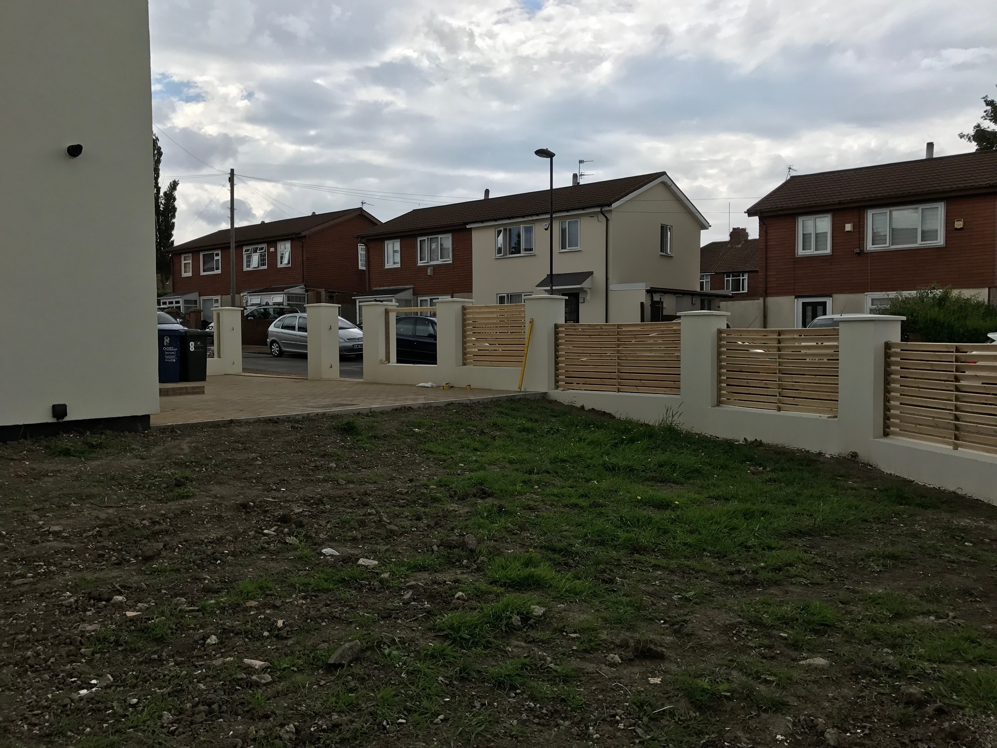 Nowoczesne ogrodzenie posesji z poziomych, drewnianych desek pomiędzy białymi, murowanymi słupkami. Na posesji świeżo nawieziona ziemia. W tle domy jednorodzinne.