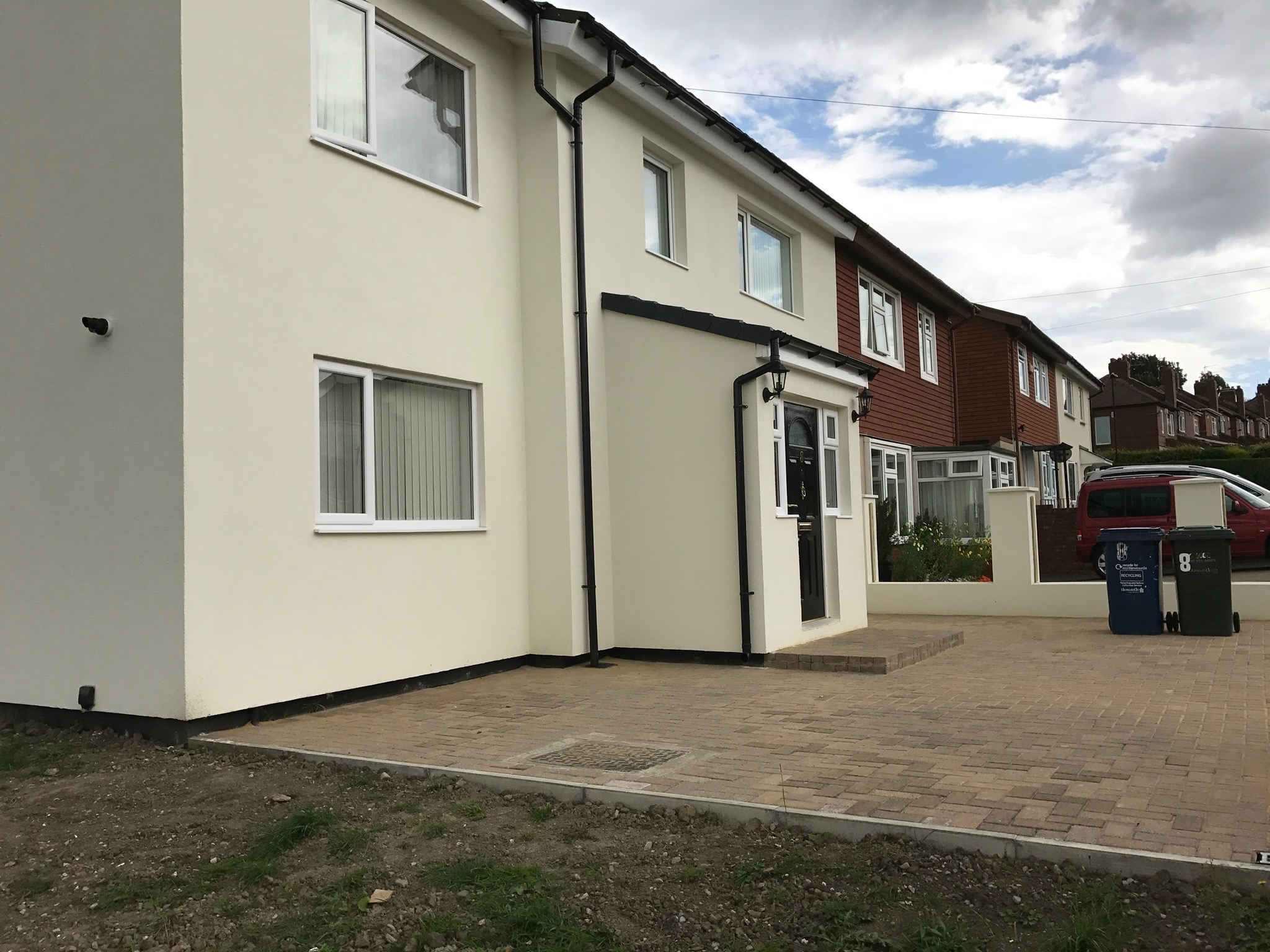 Odnowiony, dwukondygnacyjny dom jednorodzinny z jasną elewacją i czarnymi rynnami, widok z poziomu gruntu na podjazd z kostki brukowej i sąsiednie budynki.