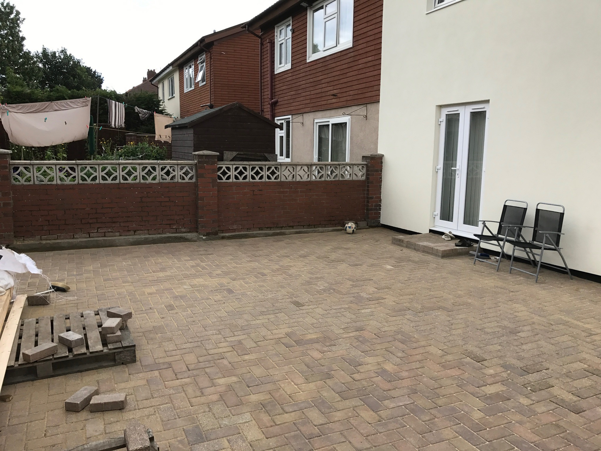 Wyłożony kostką brukową podjazd przed domem z ocieploną, jasną elewacją, widoczne przygotowane materiały budowlane na palecie i dwa krzesła przed wejściem.